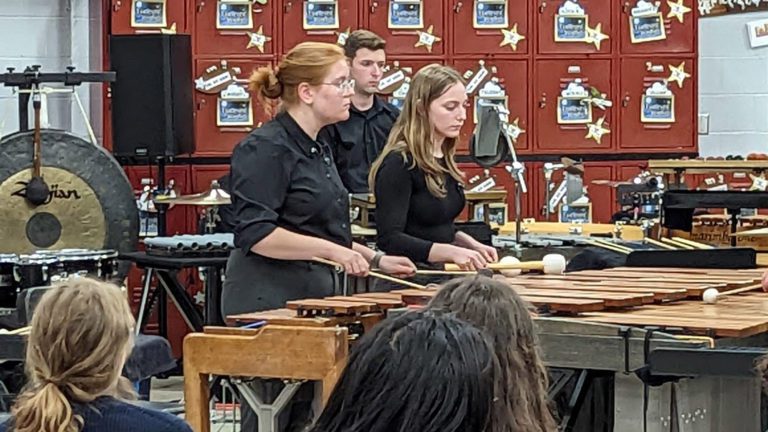 A young woman with blonde hair and glasses plays the marimba. 