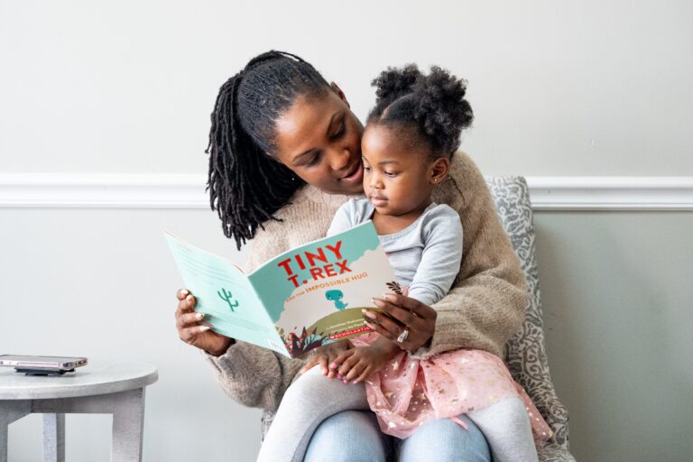 A woman sits in a chair with a child in her lap, holding a book in front of them and reading to the young girl.
