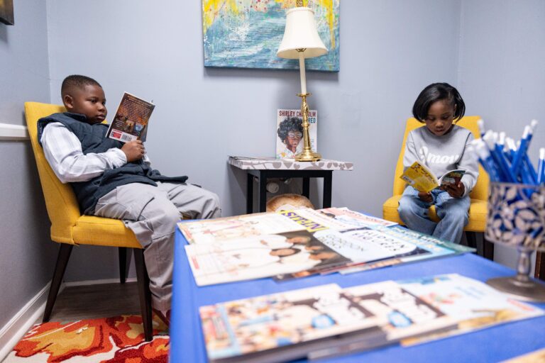 Two children sit reading in chairs in a room, with a table lined with books in front of them.