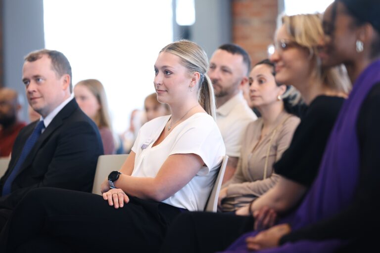 A young white woman sits in a group of people facing an off-camera speaker