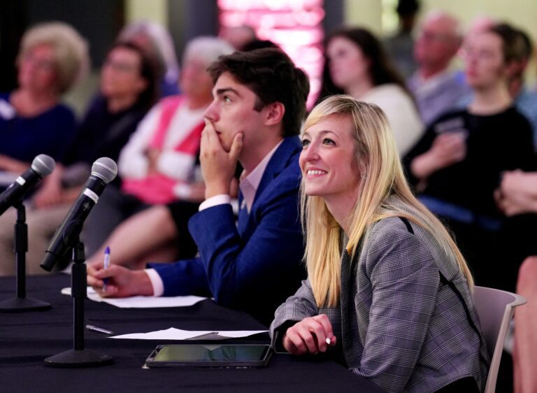 A white man and woman sit in business attire in front of two microphones on a table with an audience seated behind them