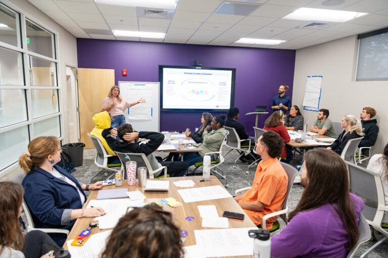 Students sitting in a classroom listening to a lecture with papers and devices on the table with a presentation screen over a purple wall.