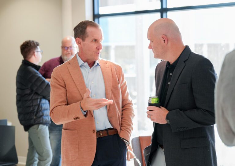 Two men in sport coats talking in front of large windows with two other people talking in the background.