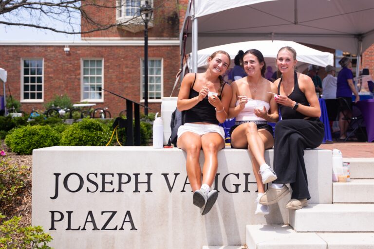 Three young women sit together on a concrete plaza sign outdoors, eating ice cream
