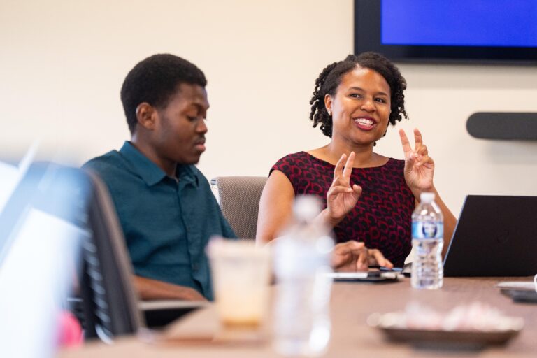 A Black man and woman sit at a long table, the woman is smiling and gesturing to people off camera as she makes a research presentation