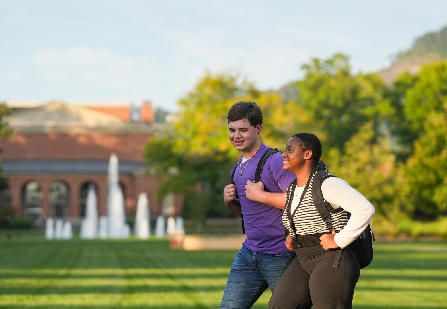 A young white man and a young Black woman walk across campus. A fountain is in the background.