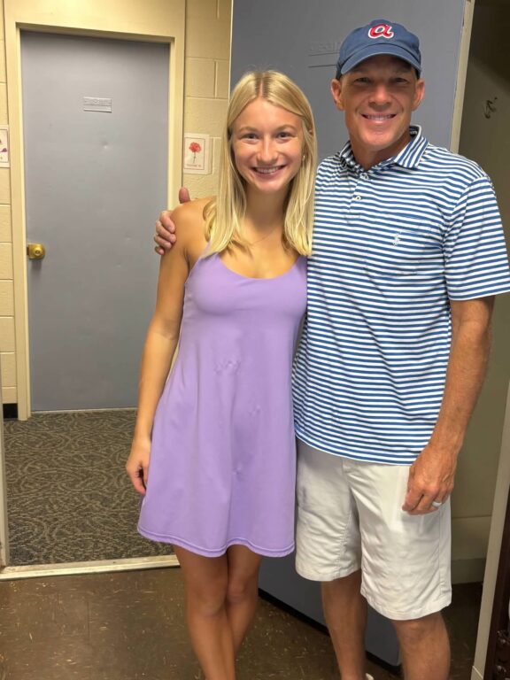 A young woman and her father pose for a photo standing in the doorway of a college dorm room.